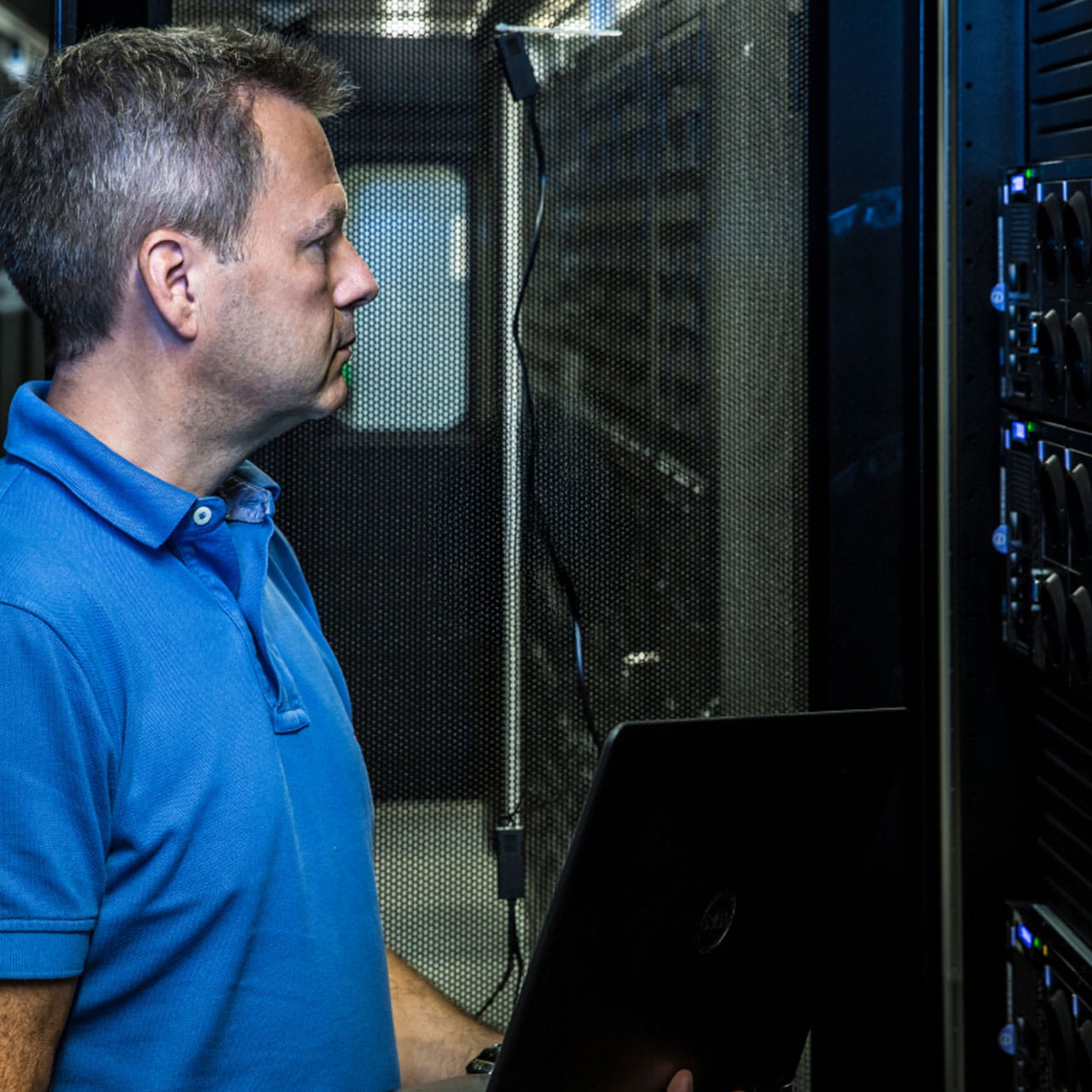 A man in a blue polo shirt looks at server racks while holding a laptop.